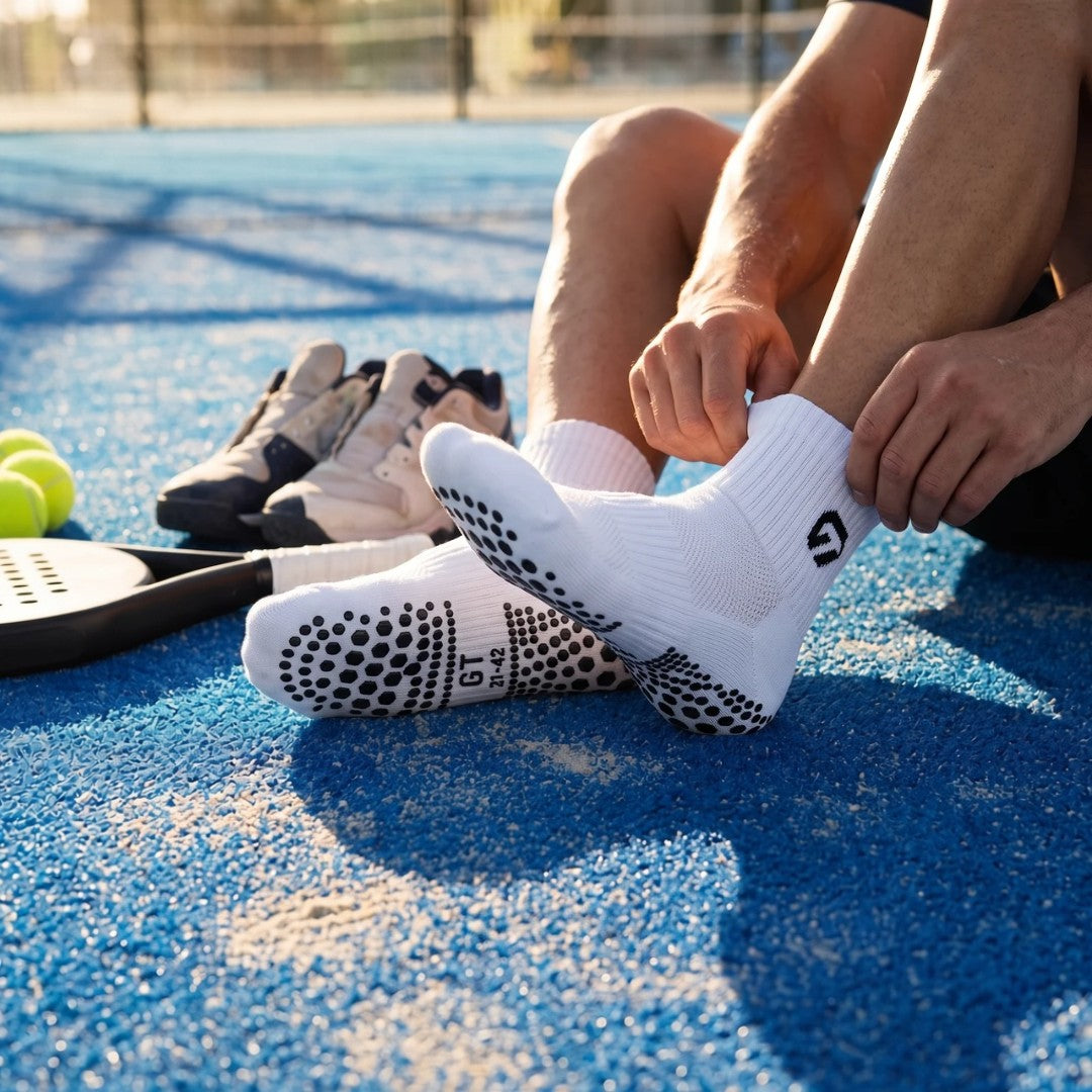Padelspeler trekt witte GT Ankle Grip Socks aan op de padelbaan, met racket, ballen en schoenen ernaast | GT Sport Selection